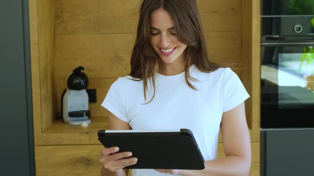 Beautiful young woman smiling while using a digital tablet in her modern kitchen, browsing social media, shopping online, or reading an e-book for a convenient and connected lifestyle