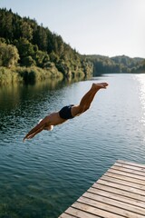 Fototapeta premium Athletic man diving from a wooden dock into a clear lake summer landscape for outdoor sport background and travel lifestyle banner