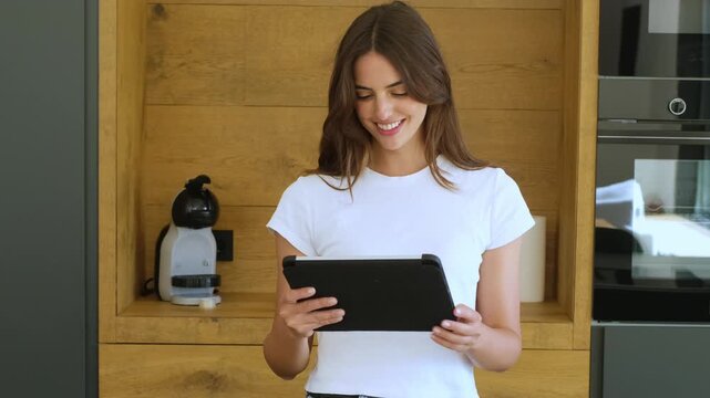 Beautiful smiling woman standing in her modern kitchen while using a digital tablet for browsing social media, reading news, or watching videos on a streaming service