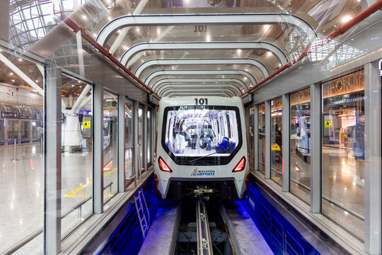 KLIA aerotrain arriving enters station through glass tunnel. Bright lights reflect off polished floors. Passengers wait calmly, eyes focused ahead.