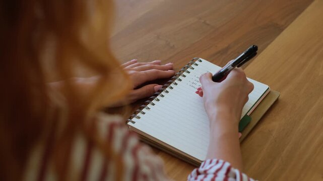Over-the-shoulder view of a redhead woman writing a to-do list in a spiral notebook, scheduling her day, and planning tasks while sitting at a wooden desk with a pen in hand