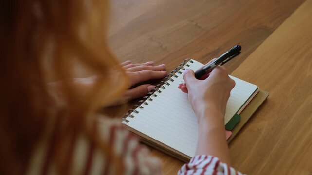 Over the shoulder view of a woman with red hair sitting at a wooden table and handwriting a to-do list in her planner, organizing her tasks and setting goals for the day