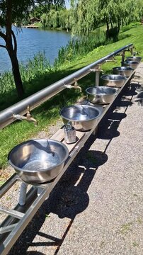 Stainless steel handwashing basins with brass taps stand in a row near a calm summer lake in the park. Concept of public hygiene, outdoor camping facilities, and summer background recreation