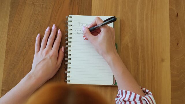 Top-down view of female hands writing a to-do list with a black pen in a spiral notebook, meticulously planning her day and setting goals on a clean wooden desk surface at home