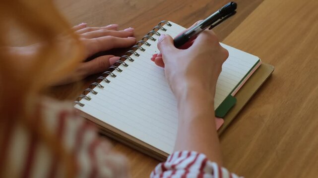 Over the shoulder view of a woman's hands writing a to-do list in a spiral notebook with a black pen, planning her daily tasks and improving her time management and productivity on a wooden desk