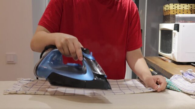 A person presses a checked kitchen towel with a steam iron on an ironing board in a home kitchen.