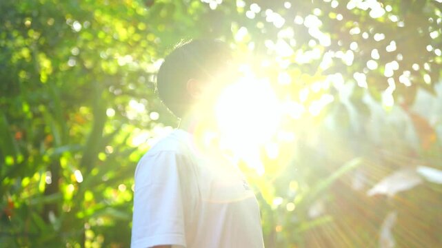 Young man shields eyes from bright sunbeams. Green foliage frames golden sunlight. He stands in natural, vibrant outdoor setting. Peaceful moment captures warmth and serenity