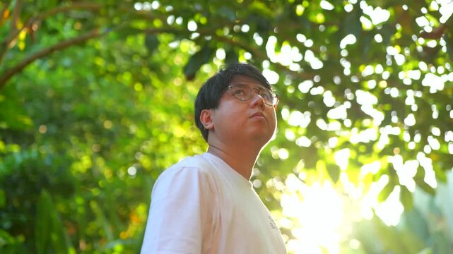 Young man shields eyes from bright sunbeams. Green foliage frames golden sunlight. He stands in natural, vibrant outdoor setting. Peaceful moment captures warmth and serenity