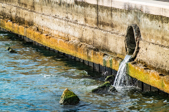 Water flows from a stone drain into a river in a rural area during daylight hours near a natural landscape with grass and rocks
