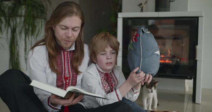 Man Reads Book with Kid Boy Together on Floor Near Fireplace. Happy Family in Ukrainian Traditional Attire. 4K 10-bit
