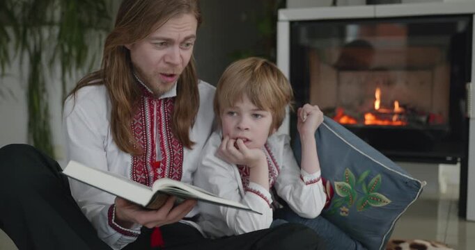 Man Reads Book with Kid Boy Together on Floor Near Fireplace. Happy Family in Ukrainian Traditional Attire. 4K 10-bit