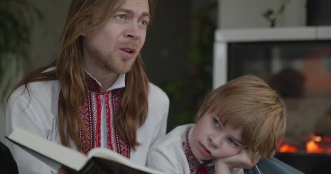 Man Reads Book with Kid Boy Together on Floor Near Fireplace. Happy Family in Ukrainian Traditional Attire. 4K 10-bit