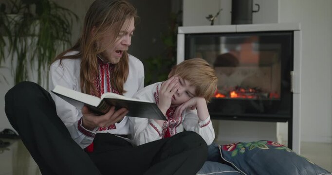 Man Reads Book with Kid Boy Together on Floor Near Fireplace. Happy Family in Ukrainian Traditional Attire. 4K 10-bit