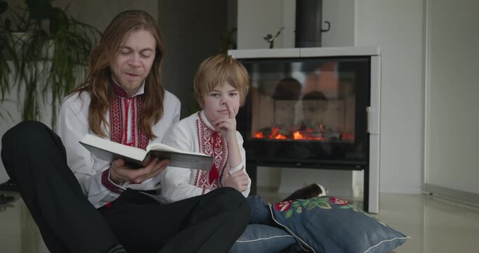 Man Reads Book with Kid Boy Together on Floor Near Fireplace. Happy Family in Ukrainian Traditional Attire. 4K 10-bit