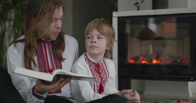 Man Reads Book with Kid Boy Together on Floor Near Fireplace. Happy Family in Ukrainian Traditional Attire. 4K 10-bit