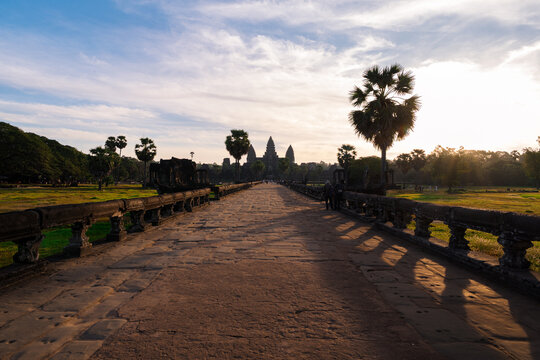 View of the historic Angkor Wat temple complex at sunset with a stone causeway leading to the main towers and palm trees in Krong Siem Reap, Siem Reap Province, Cambodia.