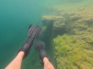 Point of view of a snorkeler wearing black flippers swimming in clear turquoise water of Rummu freshwater quarry lake near submerged limestone structures. © Dmitri