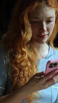 Beautiful young woman with long curly red hair using her mobile phone in the morning. Peaceful lady in pajamas enjoying her day off, checking social media and texting on her smartphone