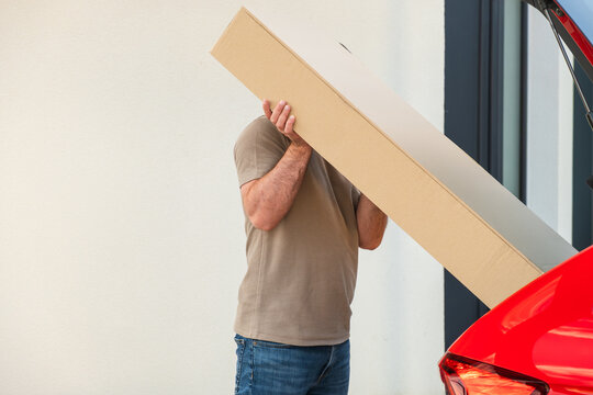 Carrying a long cardboard package and loading it into the trunk of a car in a residential area.
