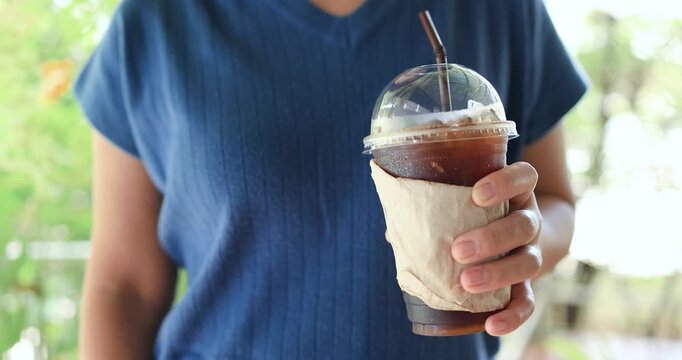Woman holding an iced Americano black coffee in a takeaway plastic cup with a natural green background. Refreshing cafe lifestyle, summer beverage and relaxation concept