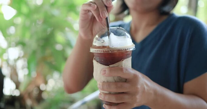 Woman holding an iced Americano black coffee in a takeaway plastic cup with a natural green background. Refreshing cafe lifestyle, summer beverage and relaxation concept