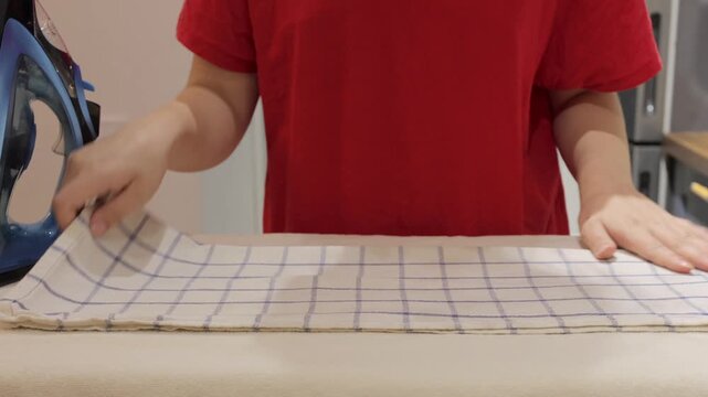 A woman folds a checked kitchen towel after ironing and smooths it on an ironing board in a home kitchen.