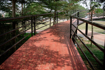 Fototapeta premium Red pedestrian bridge walkway winding through green Terminalia mantaly trees in an urban park.