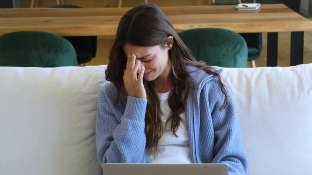 Exhausted young woman suffering from a headache while working remotely on her laptop from the couch, feeling tired and stressed due to overwork, eye strain, and professional pressure