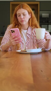 Beautiful redhead woman with long hair wearing pajamas checking social media on her phone while enjoying a cup of coffee and breakfast at home in her modern kitchen during the morning