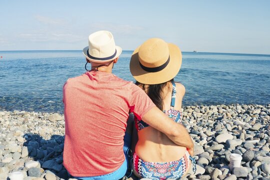 Caucasian Man and woman in summer hats embracing while sitting on sunny pebble beach facing sea horizon, concept of romance, wellness, wellbeing relaxation, travel and seaside holiday