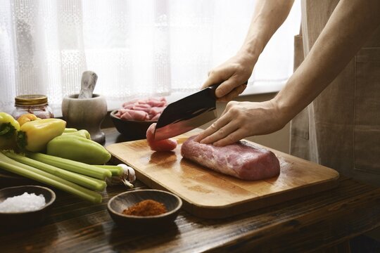 A person slices pork on a wooden cutting board surrounded by colorful vegetables and spices. Bright daylight streams through the window, illuminating the kitchen atmosphere