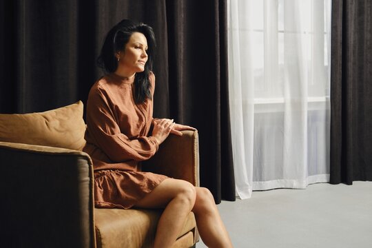 A woman sits on a comfortable couch, gazing out contemplatively. She is dressed in a relaxed outfit and surrounded by softly lit curtains. The atmosphere is serene and introspective