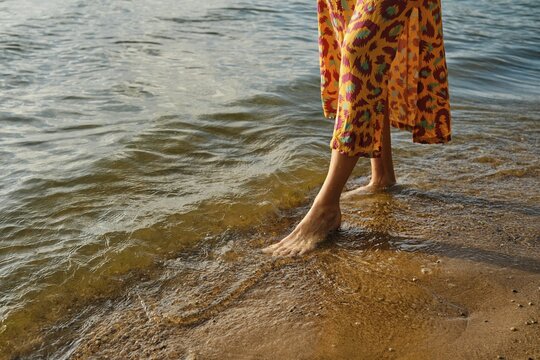 Unrecognizable woman walks barefoot along a sandy beach as waves gently lap at her feet. She wears a vibrant dress with a bold pattern, and the sun sets, casting warm hues on the water