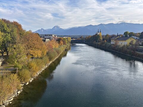 River through a city, autumn trees and mountains in the background under a clear sky, Drau, Drau cycle path, Carinthia, near Villach, Austria