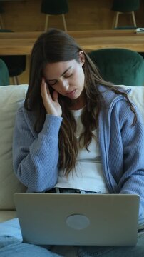 Young woman sitting on a sofa feeling tired and stressed while working on her laptop, massaging her temples to relieve a severe headache caused by overworking and intense concentration