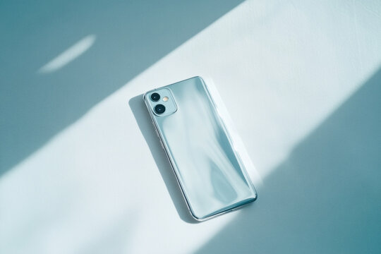 White smartphone lies on a soft lit minimalist background with diagonal shadows, highlighting glossy surface and dual camera. Composition leaves ample space for text or product overlay.
