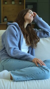 Young beautiful woman resting on the sofa in her living room, smiling peacefully and daydreaming while looking out the window with a thoughtful and serene expression on her face