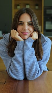 Charming young woman with a genuine smile resting her chin on her hands while sitting at a wooden table, looking at the camera with a happy and relaxed expression indoors