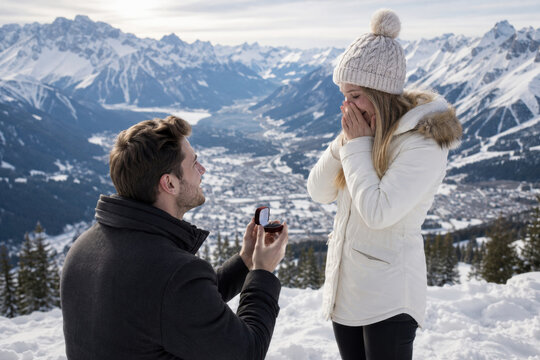 A man kneels in the snow, proposing with a ring to a surprised woman in a winter coat. Scenic mountain backdrop and emotional moment.