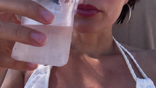 Close up of young woman sunbathing on sun lounger and drinking cold water from plastic glass to prevent dehydration during hot summer day on beach