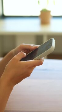 Close-up view of female hands holding a mobile phone, actively scrolling, swiping, and typing on the touch screen while sitting at a wooden table in a brightly lit room with a plant