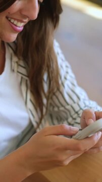 Close-up of a happy young woman with long brown hair, sitting at a table and smiling while texting, chatting, and scrolling on her smartphone, enjoying social media communication