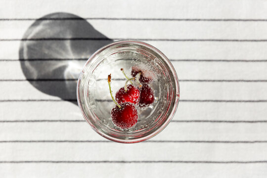 Verre transparent rempli d'eau p&eacute;tillante avec des cerises rouges remontant &agrave; la surface, nappe ray&eacute;e blanche et noire, bulles, vue du dessus, ambiance lumineuse et rafra&icirc;chissante pour l'&eacute;t&eacute;