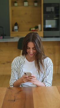 Beautiful young woman sitting at a wooden table in her modern kitchen, smiling happily while texting, scrolling, and browsing on her smartphone, enjoying online communication and social media