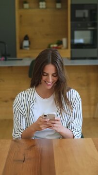 Cheerful beautiful woman sitting at a wooden table in her modern kitchen while using her smartphone for social media, chatting, and browsing, showing genuine happiness and connectivity