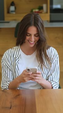 Beautiful young woman sitting at a wooden table in her modern home, smiling while using her smartphone for texting, browsing social media, or connecting with friends online