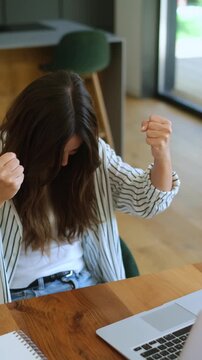 Joyful young woman clenching her fists and celebrating a victory while sitting at her desk with a laptop, expressing excitement and accomplishment after receiving good news online
