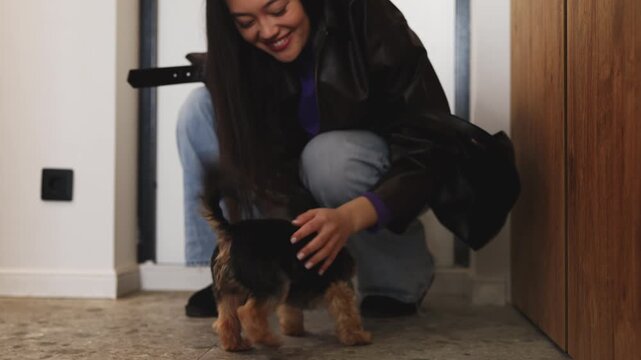 Smiling Asian woman returning home and greeting her cute puppy in the hallway