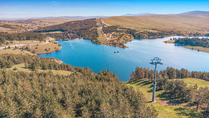 Zlatibor Ribnicko jezero reservoir with gondola cabins traversing over pine forests and hills, offering a beautiful natural landscape © EdNurg
