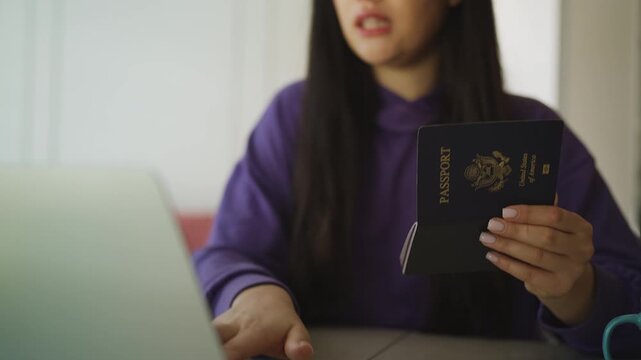 Happy Asian woman holding an American passport while booking a hotel on a laptop